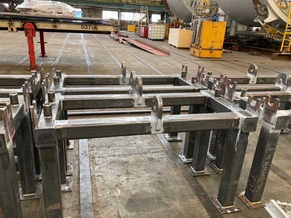 Row of fabricated steel support stands with clevis lugs on the floor of a fabrication workshop