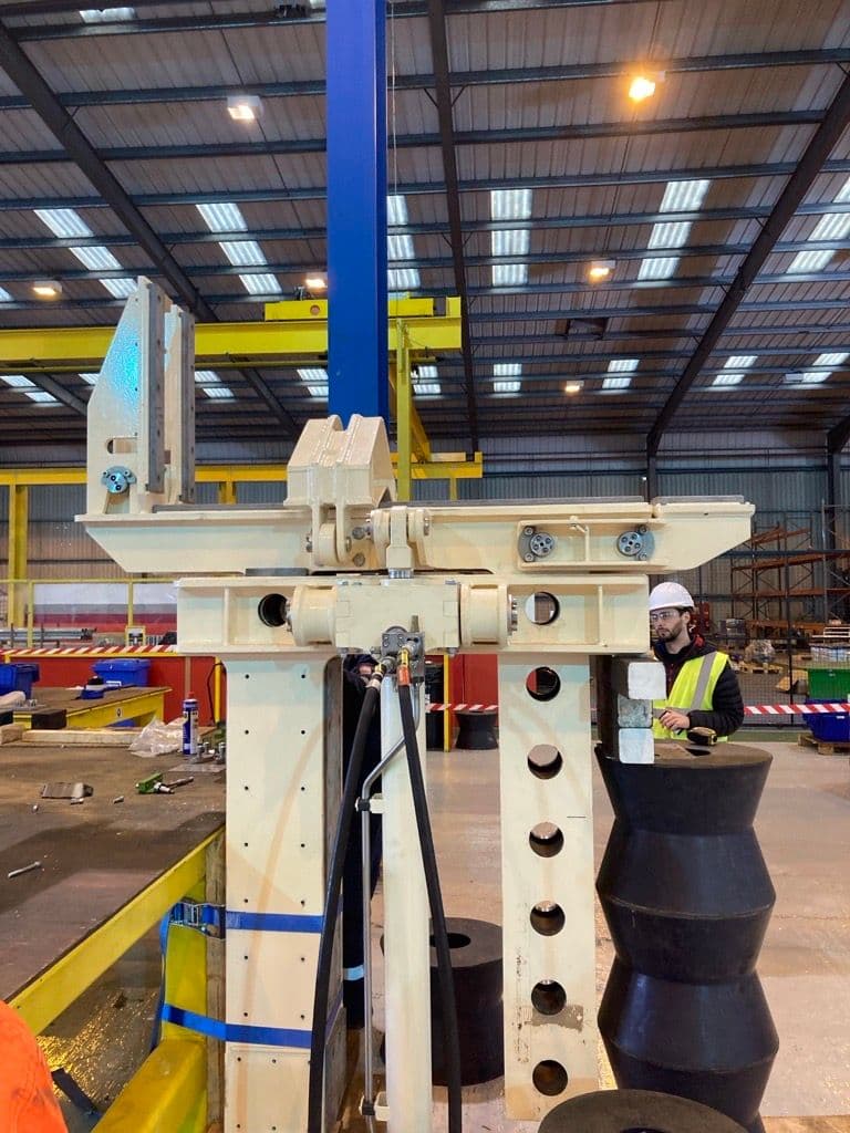 Bespoke fabricated mechanism with hydraulic hoses under assembly in a workshop, watched by an engineer in a hard hat