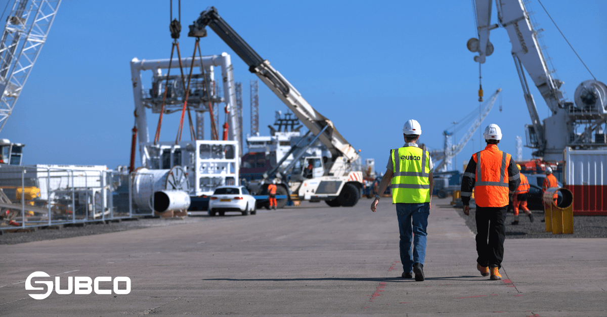 Two Subco engineers in hi-vis walk the quayside at the Port of Blyth as cranes lift offshore energy equipment behind them
