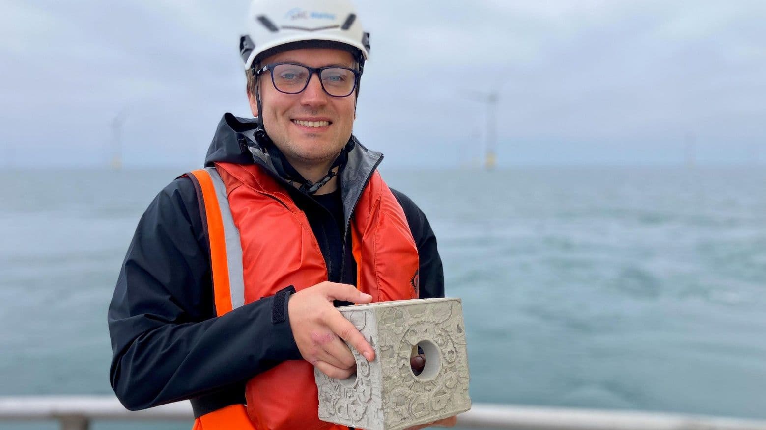 Man in a hard hat and life vest holding a holey concrete reef cube at sea, with offshore wind turbines behind